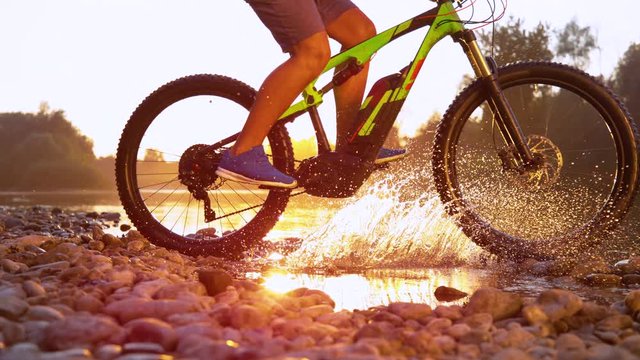 SLOW MOTION TIME REMAP SUN FLARE CLOSE UP: Unrecognizable sportsman riding a mountain bike in a shallow stream running through tranquil nature at sunset. Mountain biker splashing refreshing water.