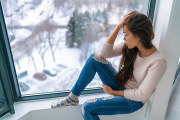 Winter depressed sad girl lonely by home window looking at cold weather upset unhappy. Bad feelings stress, anxiety, grief, emotions. Asian woman portrait.