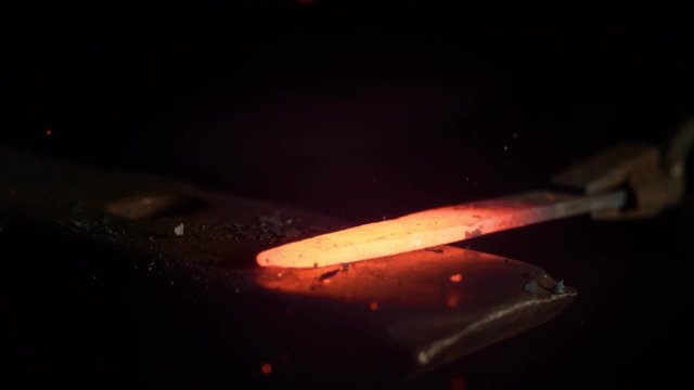 SUPER SLOW MOTION, MACRO, DOF: Red hot piece of metal is being held by tongs and struck by a big hammer. Glowing particles flying away from a knife blade being manufactured by experienced bladesmith.