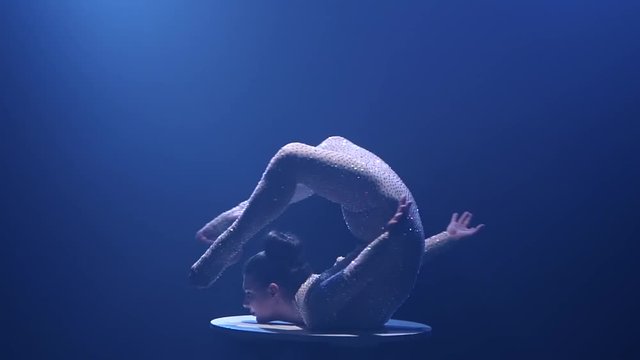 Acrobat girl spins on a table standing upside down. Smoke blue background. Slow motion