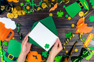 Woman hand puts blank postcard with green leprechaun lucky clover trefoil into green envelope on wooden background with props: green hat, gold coins gifts and glass of beer. Saint Patrick Day concept