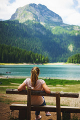 Naklejka na ściany i meble Tourist girl enjoys the magical view sitting on a wooden bench on the shore of a turquoise lake in the mountains. 