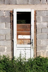 Fototapeta na wymiar Destroyed dilapidated white wooden doors with cracked paint and boards without glass mounted on brick wall of abandoned house with high uncut grass and other plants in front