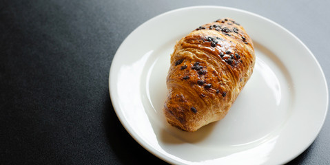 Morning Breakfast-crispy croissant with chocolate drops on a white plate stands on a black background.