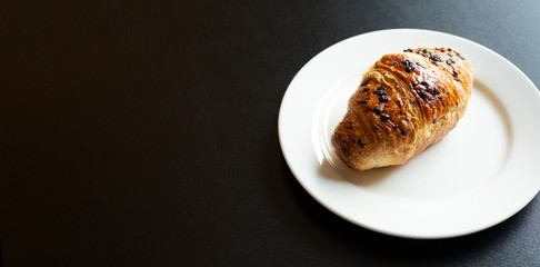 Morning Breakfast-crispy croissant with chocolate drops on a white plate stands on a black background.