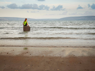 Fototapeta na wymiar Girl in wetsuit going into water with her small serf board. Mountains in the background, calm water. Concept outdoor activities, active live style.