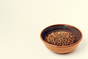 Dry raw buckwheat grains in brown ceramic bowl on white wooden background.