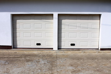 Obraz na płótnie Canvas Two modern white rollup garage doors with cracked concrete driveway mounted on white wall on warm sunny day