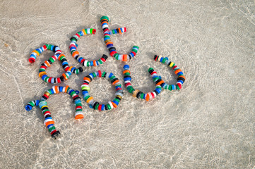 Colorful plastic bottle caps spell out 'act now' on a sandy beach with lapping waves, a reminder...