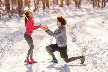 Runners on the snow. Man kneeling and giving to woman big snowball while woman laughing. Wintertime, healthy lifestyle concept.