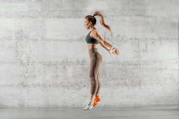 Fototapeta premium Side view of muscular focused brunette with ponytail and in sportswear jumping in front of brick wall in gym.