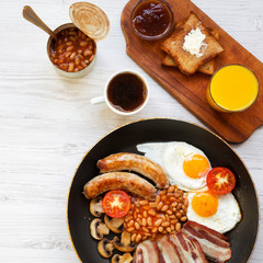 Full English breakfast in a pan with fried eggs, bacon, sausages, beans and toasts on white wooden table, overhead view. Flat lay. From above.