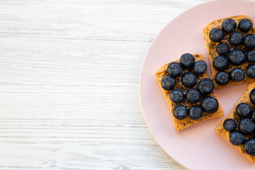 Top view, vegan toasts with peanut butter, blueberries and chia seeds on a pink plate over white wooden background. Flat lay, from above. Copy space.