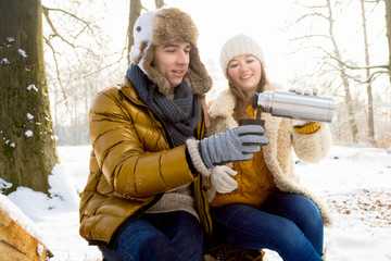 Glückliches Paar im winterlichen Wald sitzt auf einem Baumstamm und trinkt Tee