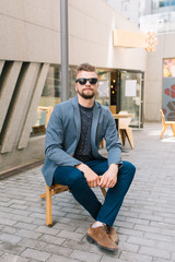 Vertical photo of handsome guy sitting on chair outdoor on cafe background. He wears gray jacket, jeans, brown shoes, sunglasses. He is looking to the camera.