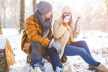 Glückliches Paar im winterlichen Wald sitzt auf einem Baumstamm und trinkt Tee
