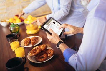 Aged couple busy look at digital tablet while having delicious breakfast at home kitchen