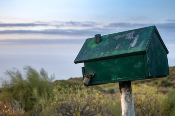 Old green wooden mailbox closed on the lock, standing in the field against the blue sky and clouds