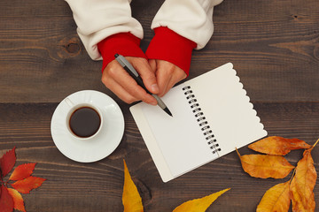 Hands of man with a pen and notebook at a wooden table with a cup of espresso and autumn leaves