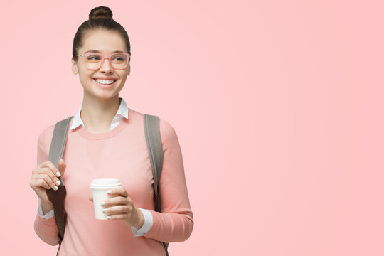 Young European Woman Student Wearing Glasses And Dressed Casually And With Backpack Behind, Holding Takeaway Coffee Cup And Smiling, Isolated On Pink Background