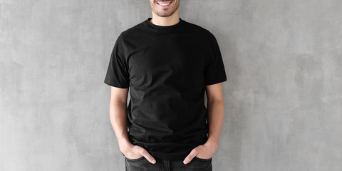 Attractive young man with poses in blank black cotton t-shirt, standing against gray textured wall