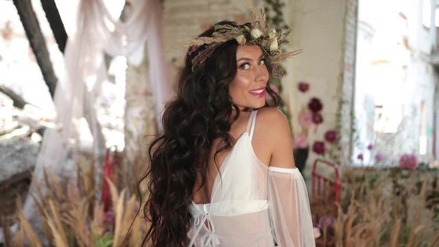 Conceptual shooting. 4k. Woman with long dark hair dressed like a nymph sits on the old bed among blooming pink flowers and hau and smiles looking straight in the camera