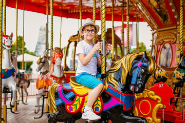 Blonde boy in the straw hat and big glasses riding colorful horse in the merry-go-round carousel in...
