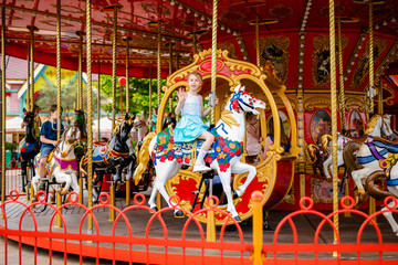 Blonde girl with two braids in white and blue dress riding colorful horse in the merry-go-round carousel in the entertainment park. Russia, Sochi, June 2018