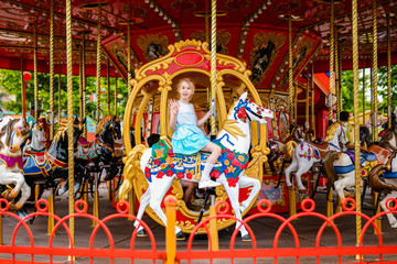Obraz na płótnie Canvas Blonde girl with two braids in white and blue dress riding colorful horse in the merry-go-round carousel in the entertainment park