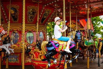 Blonde boy in the straw hat and big glasses riding colorful horse in the merry-go-round carousel in the entertainment park