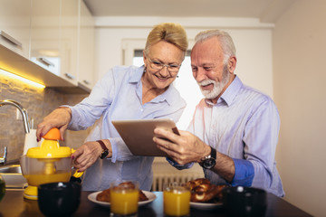 Fototapeta na wymiar Aged couple busy look at digital tablet while having delicious breakfast at home kitchen
