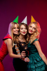 Portrait of three amazing, cute girls in fashion dresses and party caps, holding sparkler or Bengal light celebrating the New year, Christmas or Birthday party, standing over colorful background