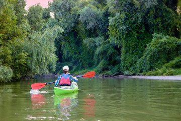 Woman paddle green kayak. Kayaking near overgrown shore of green thick thickets, wild grapes and driftwood on the banks of river at summer. Concept for adventure, travel, action, lifestyle.