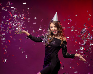 Event, party, Birthday, Christmas or New Year celebrating concept. Young pretty brunette woman in black dress and birthday hat is laughing.. Colorful studio portrait with bright background. Copy space