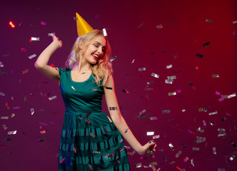 Event, party, Birthday, Christmas or New Year celebrating concept. Young pretty blond woman in green dress and birthday hat having fun with confetti falling everywhere on her. Copy space