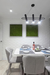 Served table with white plates glasses and bottle of wine. White kitchen interior with grey chairs and and moss on the wall in new luxury home with lights on