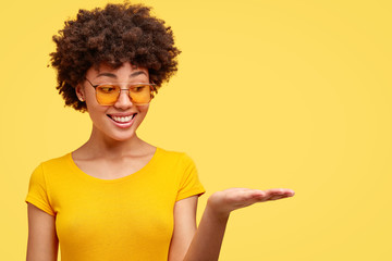 Positive African American woman with crisp hair holds copy space with one palm, has good mood, wears shades and casual t shirt, isolated over yellow background with empty space for your text