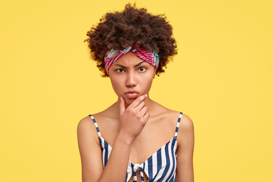 Displeased angry dark skinned young woman holds chin, claims about something bad, has sullen expression, crisp hair, wears headband, models against yellow background. Negative emotions concept