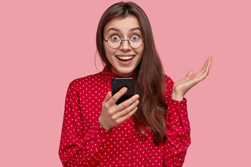Headshot of cheerful European girl holds cellular, stays in touch, sends message, happy to recieve...