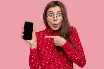 Update this application. Surprised attractive young woman points at cell phone, shocked with its...