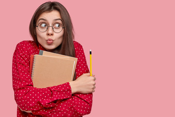 Successful female student holds notepad with pencil, creats plan for future session, makes grimace and looks aside in interest, ready for university, isolated over pink wall with copy space.