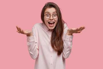 Clueless happy young European woman spreads palms, looks with hesitation and positiveness, has...