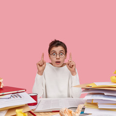 Photo of surprised young woman works at desktop, shows direction upwards, indicates with fore...