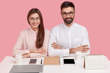 Horizontal shot of pleased self confident professional perfectionists coworkers sit next to each...