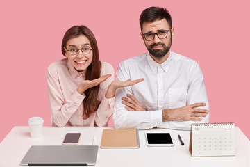 Optimistic young woman with broad smile spreads palms and shows her sad colleague, sit together at...