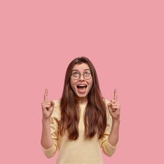 Photo of pleased emotive dark haired model opens mouth, points with both fore fingers upwards,...
