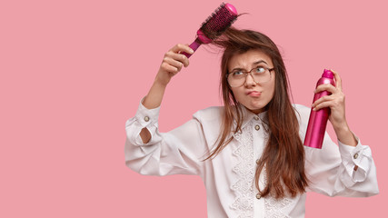 Dissatisfied young woman brushes hair, uses comb, frowns face, holds hairspray, wears white old fashionable blouse, prepares for first date, isolated over pink background with copy space aside