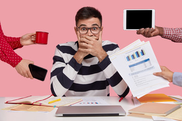 Frightened Caucasian young man covers mouth with both hands, afraids of difficulties at work, surrounded with modern gadgets, paper documents, mug of drink, writes in organizer. Work concept