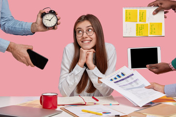 Pleased female entrepreneur keeps hands under chin, looks with gentle smile aside, poses ner...