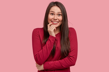 Photo of pleasant looking teen girl keeps arms partly crossed over waist, smiles broadly with intriguing expression, laughs joyfully at joke, wears red clothes, isolated over pink background